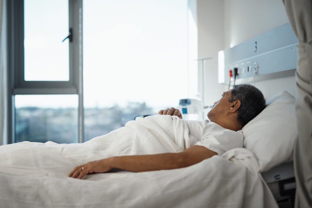 Man lying in a hospital bed, resting under a blanket near a window.