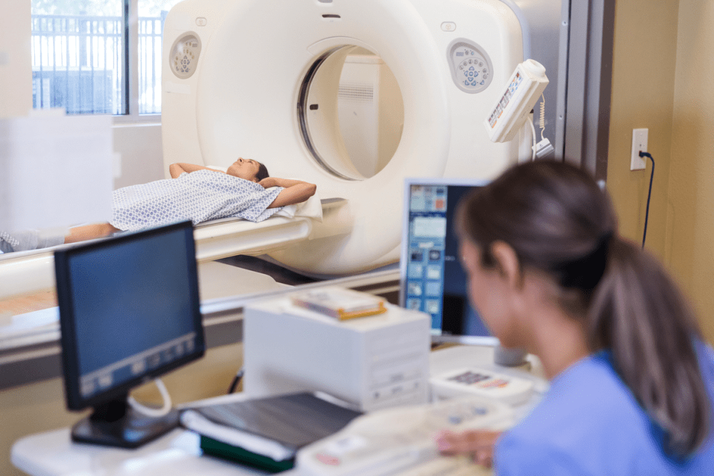 A technician sits at a desk with two monitors, preparing for a CT scan. A patient is seen in the background prepared to enter the CT Scan.