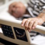 Elderly patient resting in a hospital bed, holding the bed rail with one hand.