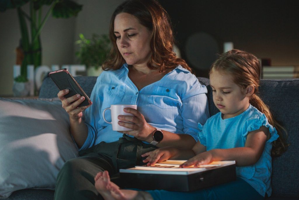 Mother with coffee mug looking at phone while daughter uses tablet