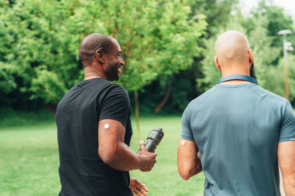 Two men wearing blood glucose monitors on their arms walk together in a park.