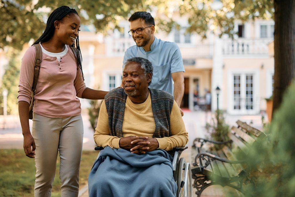 Man in wheelchair outdoors with two caregivers walking beside him.