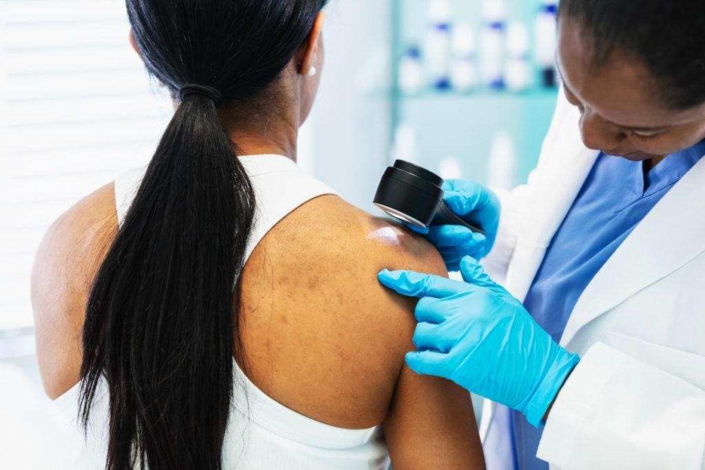 Doctor examining a skin spot on a woman’s shoulder with a dermatoscope.