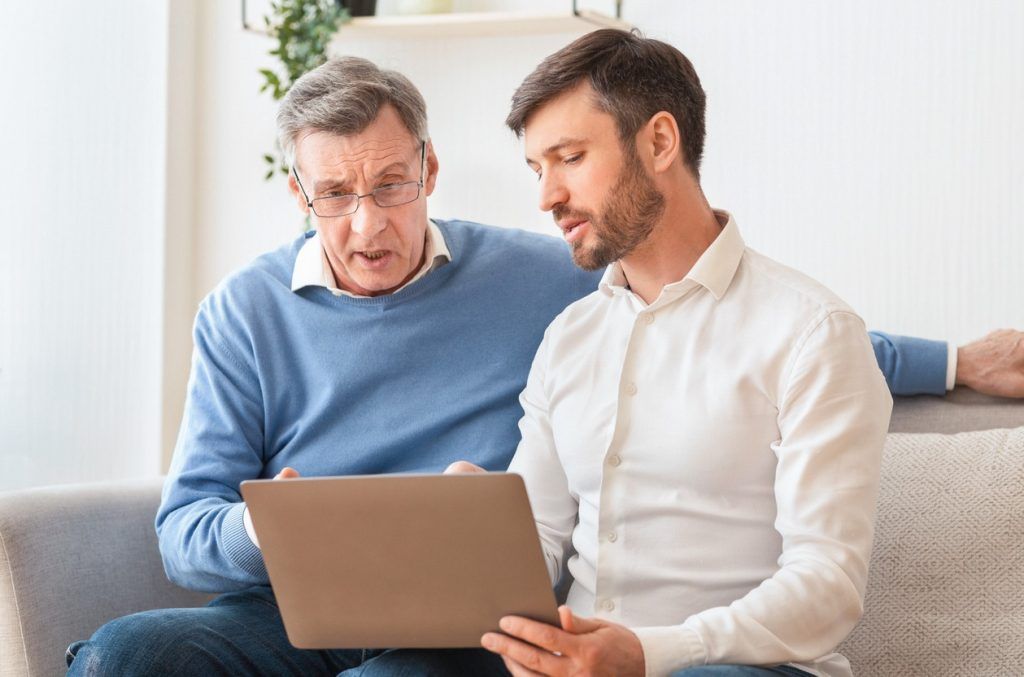 A man showing an older man something on a laptop while they both sit on the couch.