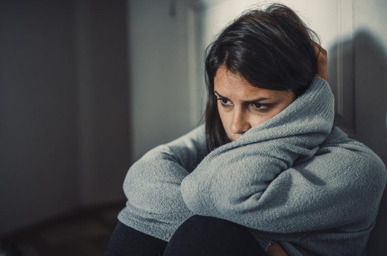 Woman sitting alone, hugging her knees, looking distressed.